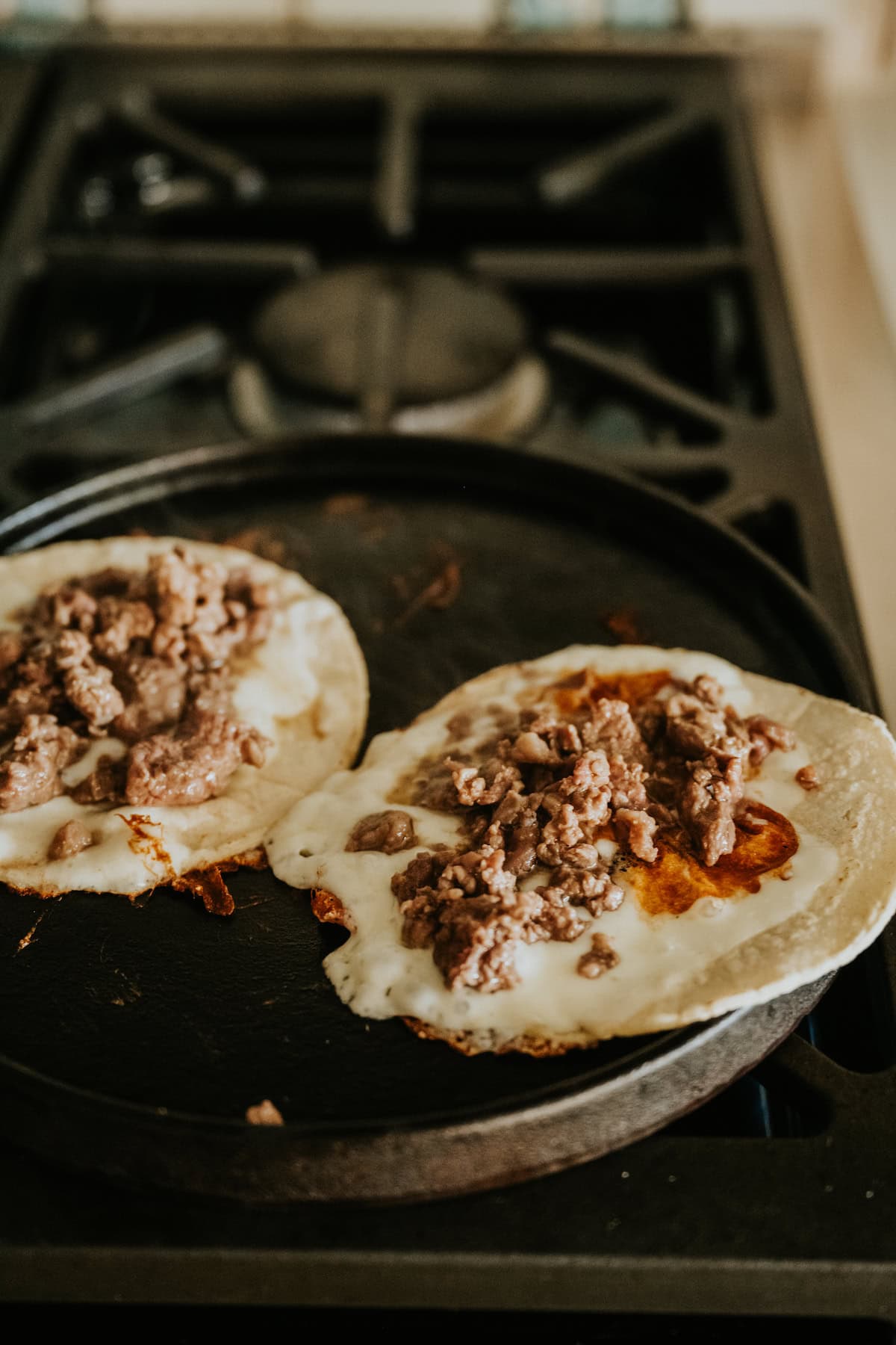 Tacos cooking on a comal with melted cheese and carne asada filling.