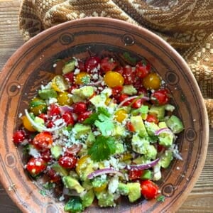 Finished Ensalada de Aguacate (Mexican avocado salad) with red and yellow cherry tomatoes, creamy avocado, red onion, cilantro, and crumbled queso fresco in a rustic clay bowl.