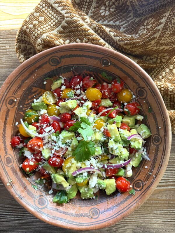 Finished Ensalada de Aguacate (Mexican avocado salad) with red and yellow cherry tomatoes, creamy avocado, red onion, cilantro, and crumbled queso fresco in a rustic clay bowl.