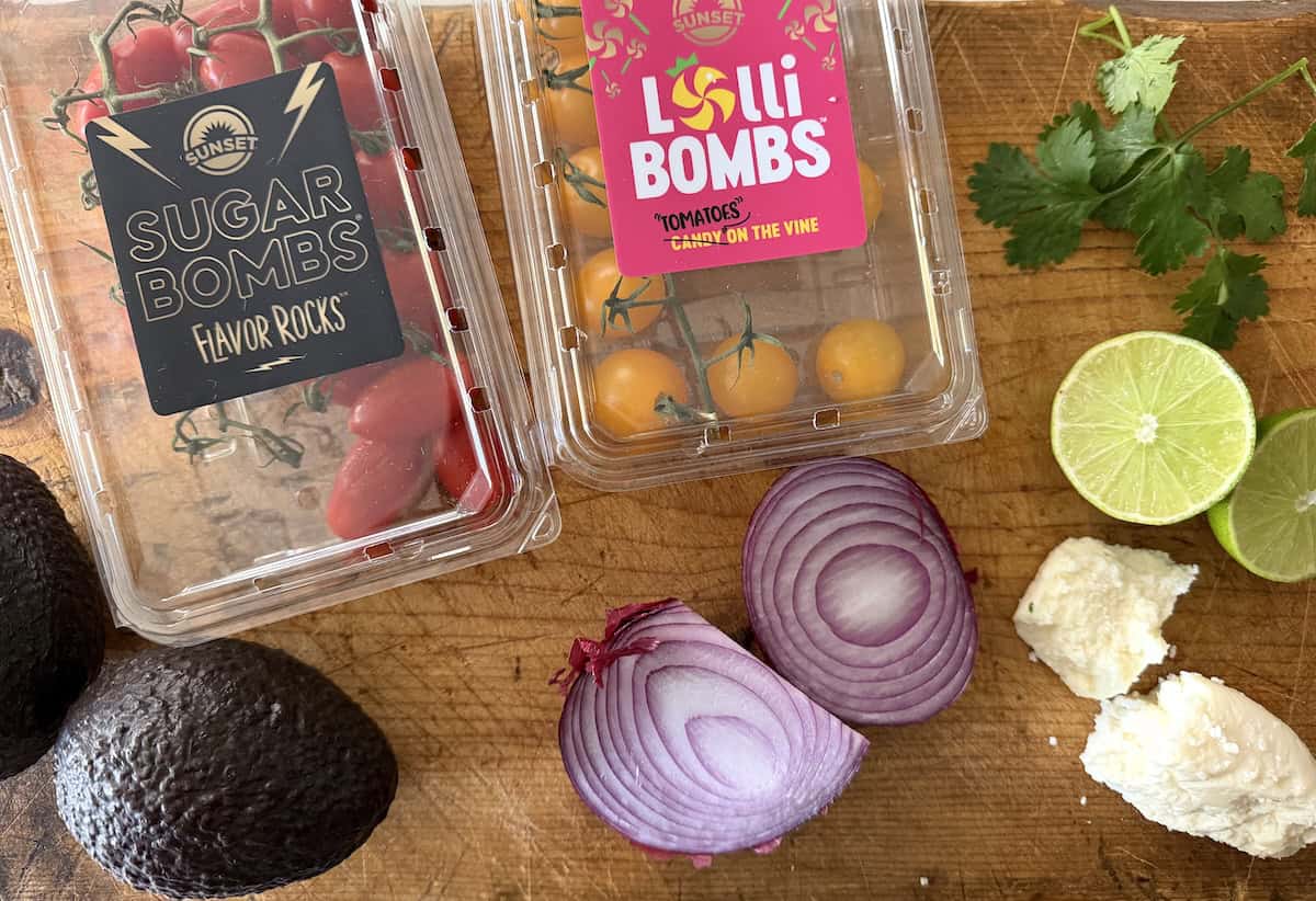Ingredients for Mexican avocado salad, including SUNSET Sugar Bombs and Lolli Bombs tomatoes, avocados, red onion, queso fresco, fresh cilantro, and lime on a wooden cutting board.