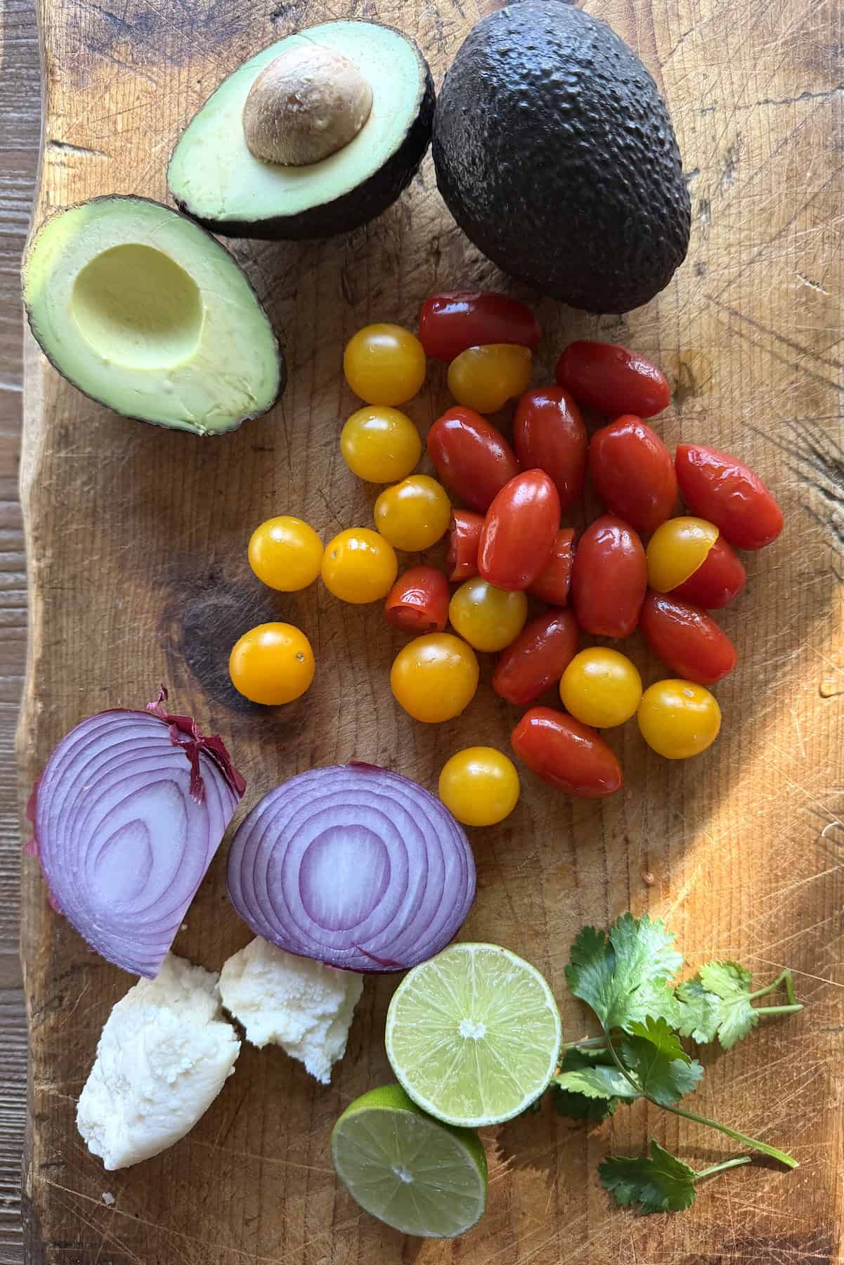 Avocados, red and yellow cherry tomatoes, sliced red onion, queso fresco, cilantro, and fresh lime on a wooden cutting board for Mexican avocado salad.