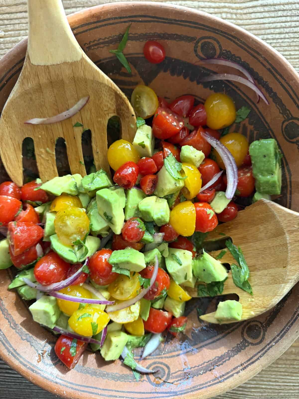 Mexican avocado salad with colorful cherry tomatoes, avocado, red onion, and cilantro being gently tossed with a wooden spoon in a clay bowl.