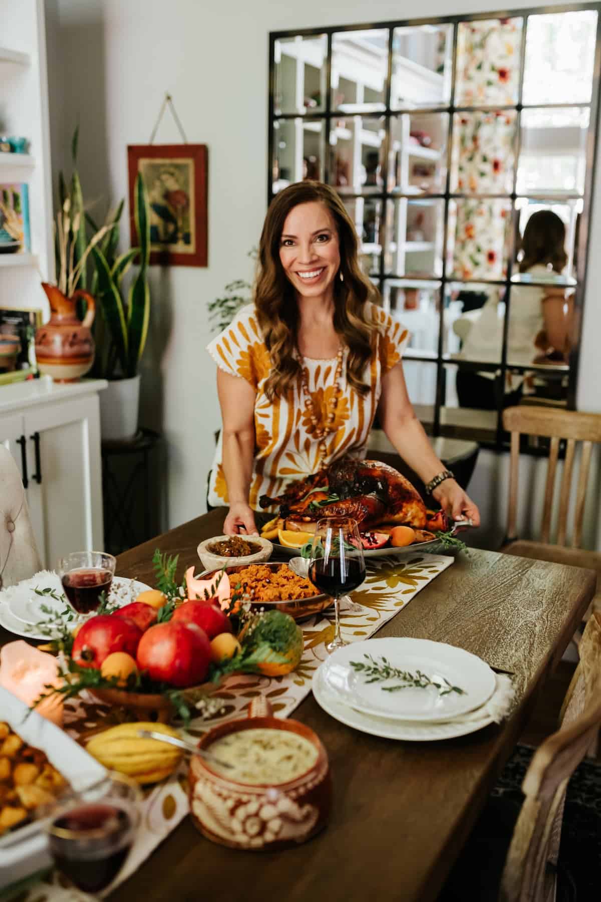 Muy Bueno cookbook author Yvette Marquez-Sharpnack standing behind a Mexican-inspired Thanksgiving table featuring Pavo Enchilado roast turkey, colorful side dishes, and festive fall decorations.